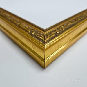 May include: Close-up of a gold picture frame with ornate detailing. The frame has a smooth, polished finish and a slightly raised edge.