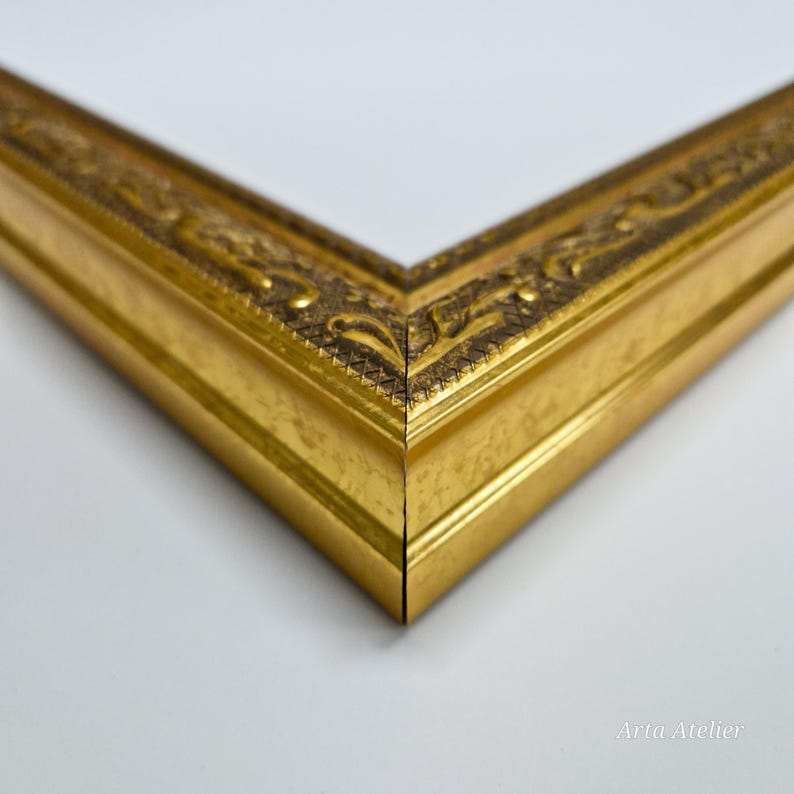 May include: Close-up of a gold picture frame with ornate detailing. The frame has a smooth, polished finish and a slightly raised edge.