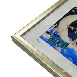 May include: A framed print of a colourful illustration with the text "Fly Me to the Moon". The print is numbered 6/200 and signed "Arta Atelier".