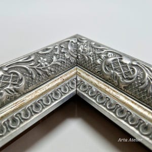 May include: Close-up of a silver picture frame with an ornate design. The frame has a raised, textured pattern of leaves and flowers. The frame is accented with a thin, gold-colored stripe.