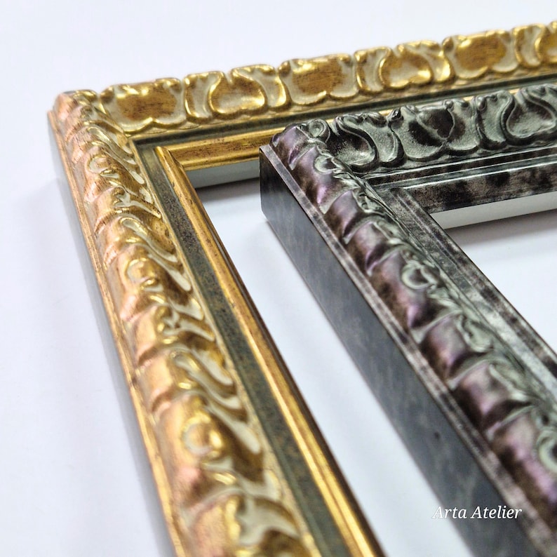 May include: Close-up of two ornate picture frames. One frame is gold with a detailed floral design, and the other is a dark gray with a similar pattern. The frames are angled, showcasing their intricate carvings and textures. The "Arta Atelier" logo is visible.