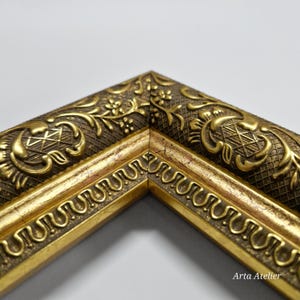 May include: Close-up of a gold picture frame with an ornate design. The frame has a raised, textured pattern with a floral motif. The frame is made of wood and has a gold finish.