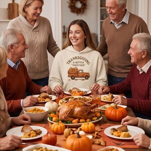 A woman wearing a cream-colored hoodie with a Thanksgiving truck graphic stands smiling at a family holiday dinner table.