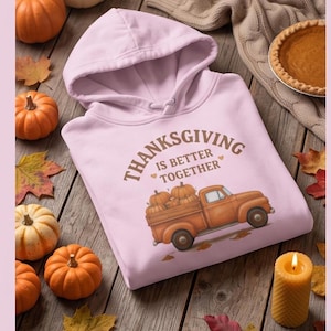 A light purple hooded sweatshirt with a Thanksgiving truck graphic is displayed on a wooden surface with small pumpkins, a candle, and a pie.
