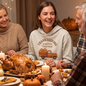 A woman in a cream-colored hoodie with a Thanksgiving truck graphic laughs with family members over a Thanksgiving dinner table with a roast turkey.
