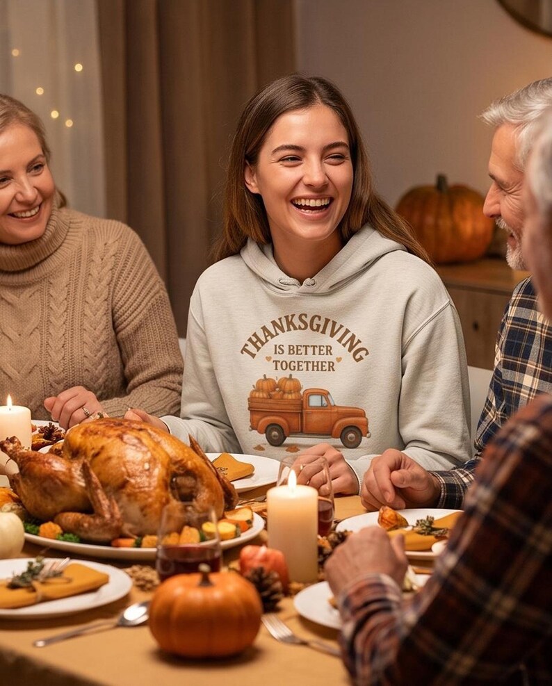 A woman in a cream-colored hoodie with a Thanksgiving truck graphic laughs with family members over a Thanksgiving dinner table with a roast turkey.