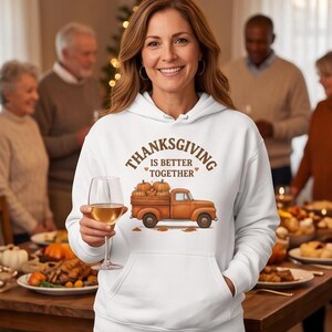 A woman wearing a white hoodie with a Thanksgiving truck graphic holds a glass of white wine at a family dinner setting.