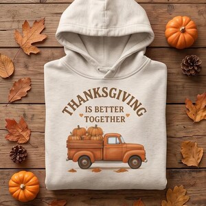 A cream-colored hooded sweatshirt with a Thanksgiving truck graphic is displayed flat on a wooden surface among pinecones, pumpkins, and autumn leaves.