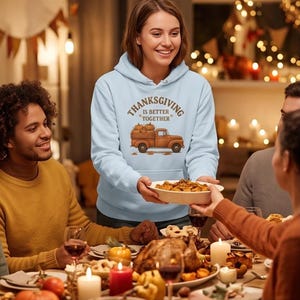 A woman wearing a light blue hoodie with a Thanksgiving truck graphic smiles as she serves food at a family dinner table.