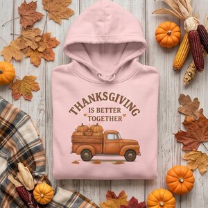 A light pink hooded sweatshirt with a Thanksgiving truck graphic is laid on wood surrounded by small pumpkins, corn cobs, and autumn leaves.