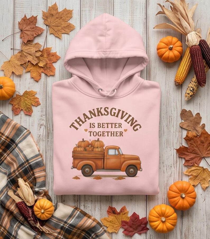 A light pink hooded sweatshirt with a Thanksgiving truck graphic is laid on wood surrounded by small pumpkins, corn cobs, and autumn leaves.