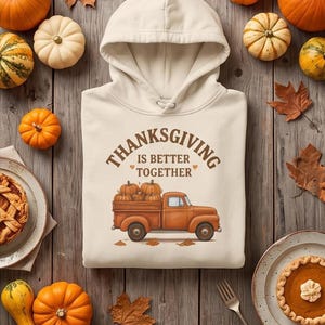 A cream-colored hoodie with a Thanksgiving truck graphic is displayed on a wooden board alongside pie, small pumpkins, and autumn leaves.