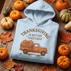 A light blue hooded sweatshirt featuring a Thanksgiving truck graphic is laid flat on a wooden surface surrounded by small pumpkins and autumn leaves.