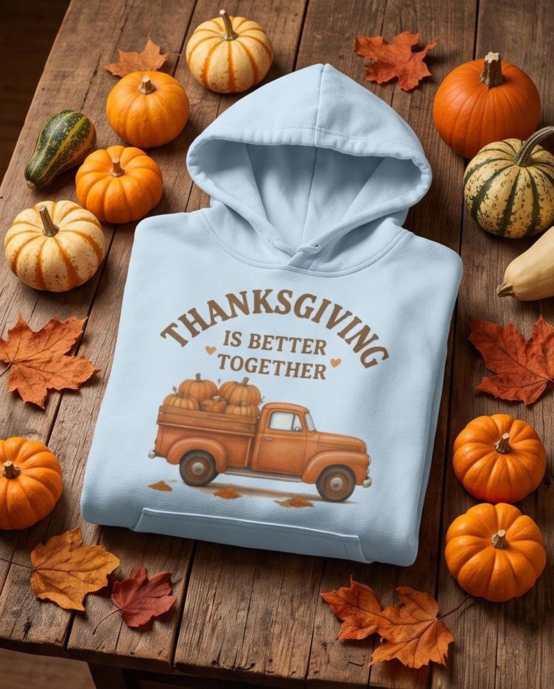A light blue hooded sweatshirt featuring a Thanksgiving truck graphic is laid flat on a wooden surface surrounded by small pumpkins and autumn leaves.