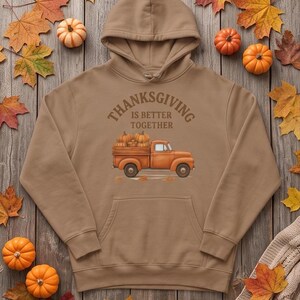 A tan hooded sweatshirt with a Thanksgiving truck graphic is laid flat on wooden boards with scattered small pumpkins and autumn leaves.