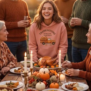 A woman wearing a pink hoodie with a Thanksgiving truck graphic smiles as she stands before a family Thanksgiving dinner table.