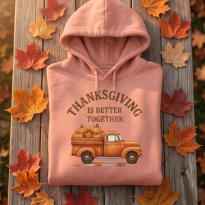 A folded pink hoodie with a Thanksgiving truck graphic is displayed on a wooden surface surrounded by scattered fall leaves.