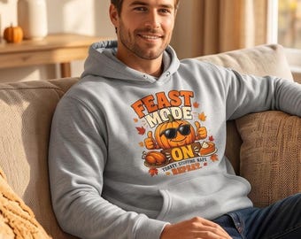 May include: A light gray hooded sweatshirt featuring a graphic print of a smiling pumpkin wearing sunglasses, giving a thumbs up. The text reads FEAST MODE ON, TURKEY STUFFING NAPS REPEAT, accompanied by illustrations of a roasted turkey and pumpkin pie.