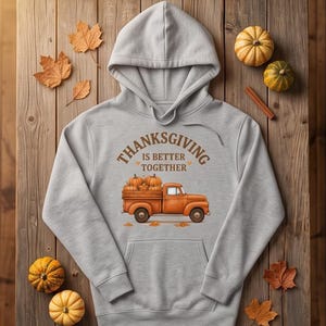 A grey hoodie with a Thanksgiving truck graphic is laid flat on a rustic wooden table with autumn leaves, small pumpkins, and cinnamon sticks.