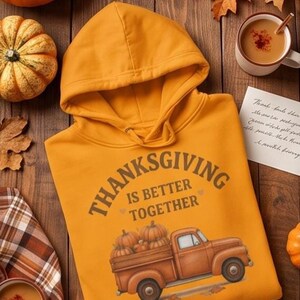 A yellow hoodie with a Thanksgiving truck graphic is arranged on a wooden surface with pumpkins, autumn leaves, mugs of cider, and a handwritten note.