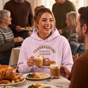 A laughing woman wearing a light purple hoodie with a Thanksgiving truck graphic holds a mug of coffee at a family dinner table.