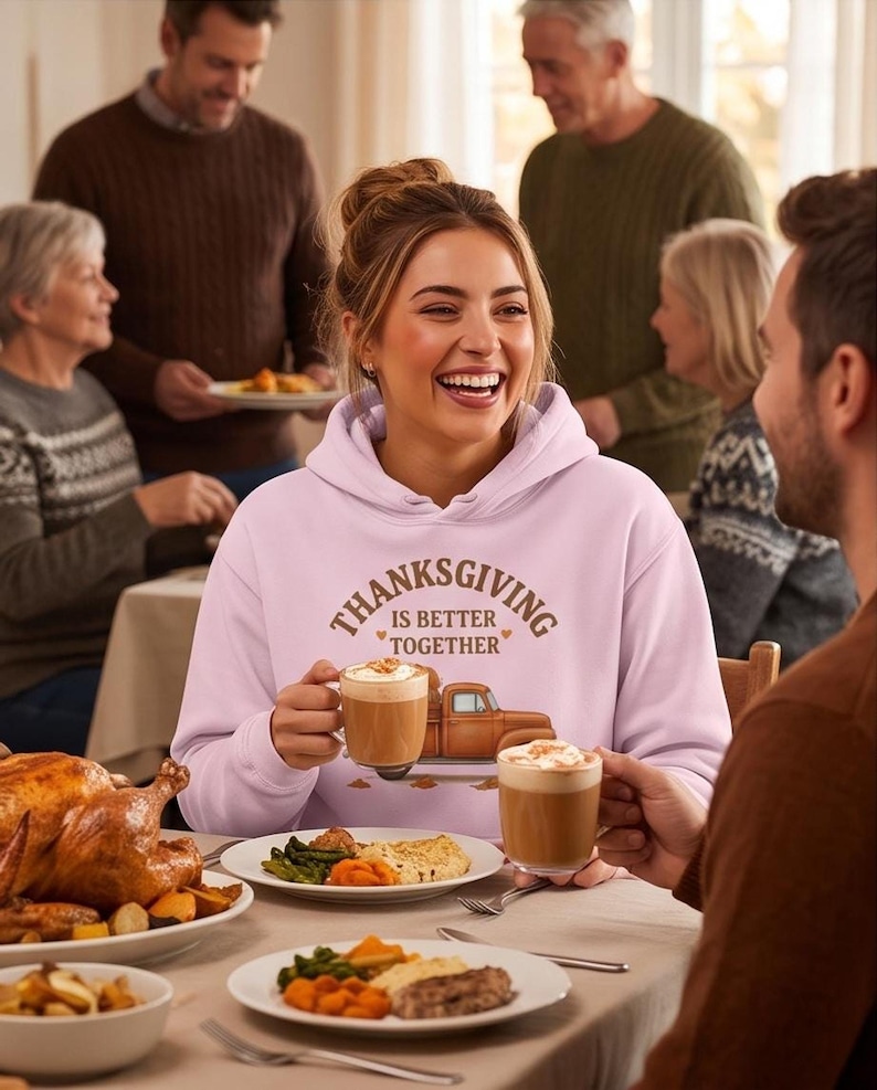 A laughing woman wearing a light purple hoodie with a Thanksgiving truck graphic holds a mug of coffee at a family dinner table.