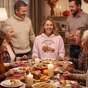 A smiling woman wearing a pink hoodie with a Thanksgiving truck graphic sits at a crowded family dinner table with lit candles and food.