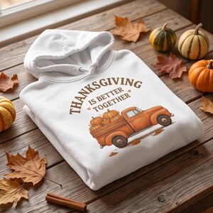A folded white hoodie with a Thanksgiving truck graphic sits on a wooden surface with scattered autumn leaves and cinnamon sticks.