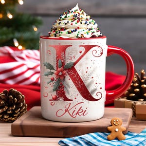 Pode incluir: Uma caneca de cerâmica branca com alça e borda vermelhas, decorada com a letra "K" vermelha e folhagens festivas. A caneca é preenchida com chantilly e granulado colorido, com o nome "Kiki" na frente.