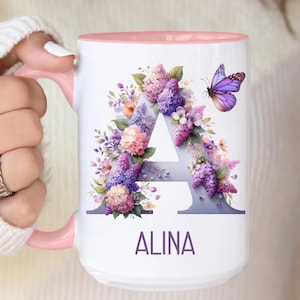 Puede incluir: Taza de cerámica blanca con interior y asa rosas. La taza presenta un diseño floral alrededor de la letra "A" en tonos morados, rosas y blancos, con una mariposa morada. El nombre "ALINA" está impreso en morado debajo.