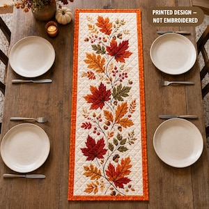 May include: A rectangular table runner with a cream-colored quilted background and a printed design of autumn leaves, berries, and acorns in shades of red, orange, and green. The runner features a bright orange border and text stating PRINTED DESIGN – NOT EMBROIDERED.
