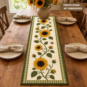 May include: A rectangular table runner with a cream-colored quilted center featuring a printed design of yellow sunflowers, green leaves, and small bees. The runner has a green and white checkered border and includes the text PRINTED DESIGN – NOT EMBROIDERED.