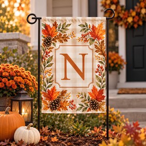 May include: A rectangular garden flag featuring a beige and white buffalo check pattern. The centre displays a large orange letter N inside a decorative border. The border is surrounded by autumn foliage, including maple leaves, pinecones, and berries.