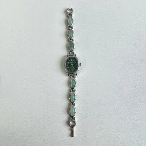 May include: Silver-toned wristwatch with a green face and matching oval-shaped green stones along the band. The watch has a rectangular face with a green dial and silver hands. The band is made of silver links with oval green stones.