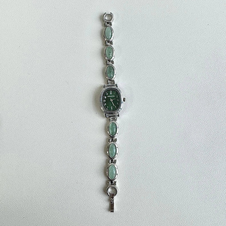 May include: Silver-toned wristwatch with a green face and matching oval-shaped green stones along the band. The watch has a rectangular face with a green dial and silver hands. The band is made of silver links with oval green stones.