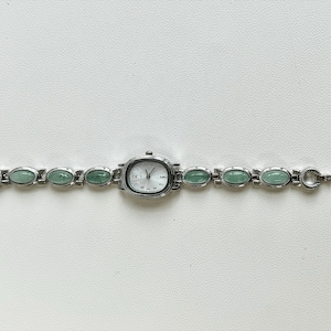 May include: A silver-toned wristwatch with an oval face and a light-colored dial. The watch band features oval, light green stones. The watch is displayed on a white surface.