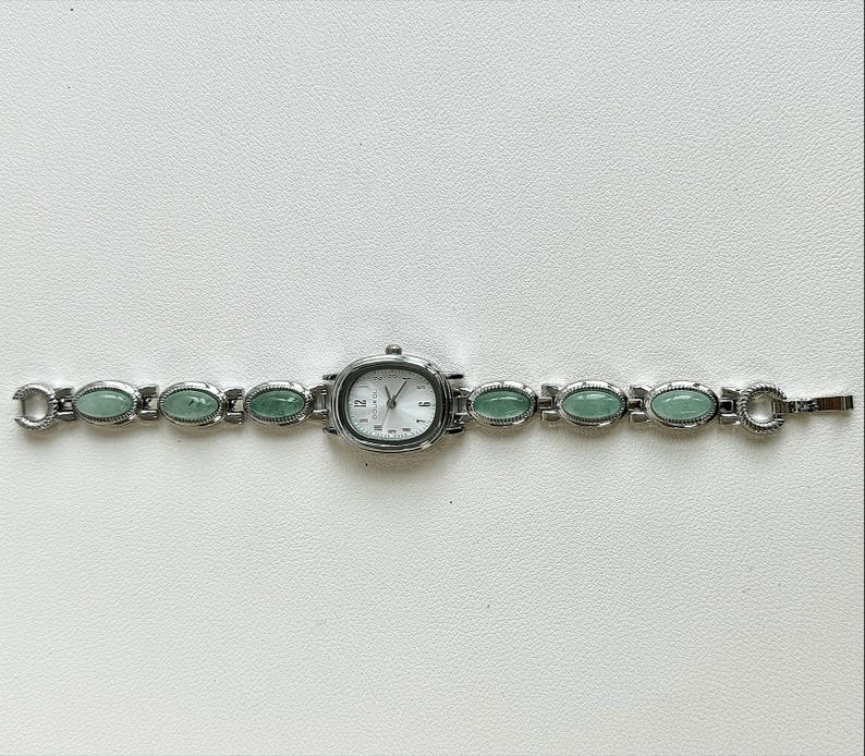 May include: A silver-toned wristwatch with an oval face and a light-colored dial. The watch band features oval, light green stones. The watch is displayed on a white surface.