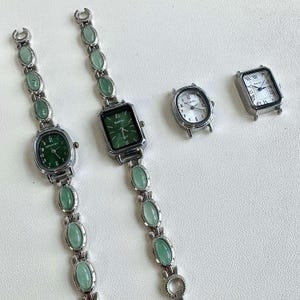 May include: Silver-toned watches with green faces and oval-shaped green stone accents. Two watches have matching stone bracelets. One watch has a white face with black numerals, and another has a green face with black numerals. The watches have a vintage aesthetic.