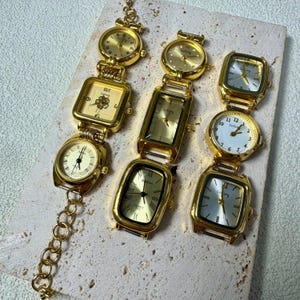May include: A collection of gold-toned watches with various face shapes, including round, square, and rectangular. Some watches have Roman numerals, while others have Arabic numerals. The watches are arranged on a light-colored surface.
