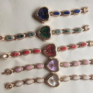 May include: Five gold-coloured heart-shaped watches with gemstone bracelets. The watch faces are blue, green, red, purple, and white. The bracelets feature oval gemstones in matching colours.