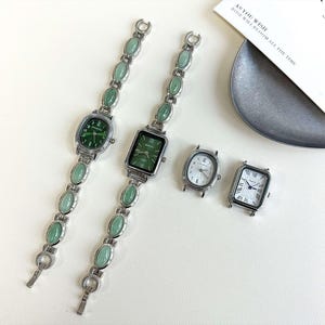May include: Several silver-toned watches with different face shapes and green or white dials. Two watches have green oval-shaped stones on the bands. One watch has a rectangular face with the word "BANSH" on the dial. The other watches have white dials.