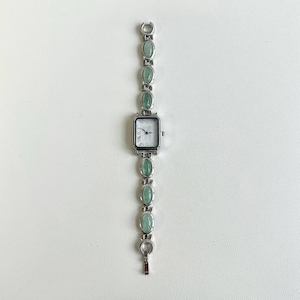 May include: A silver-toned wristwatch with a rectangular face and Roman numeral hour markers. The watch band features oval-shaped, light green stones. The watch is displayed on a white surface.