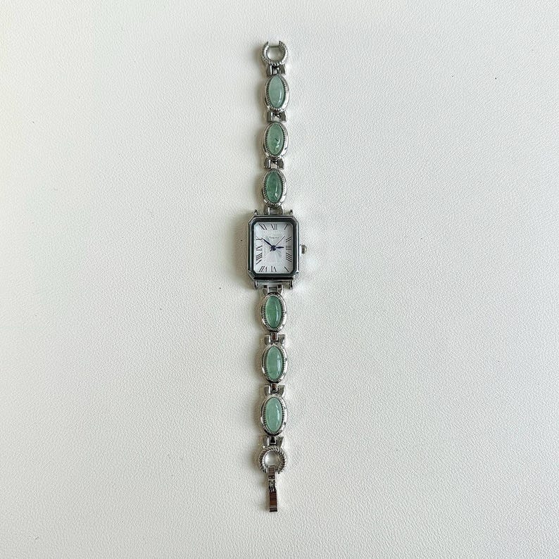 May include: A silver-toned wristwatch with a rectangular face and Roman numeral hour markers. The watch band features oval-shaped, light green stones. The watch is displayed on a white surface.