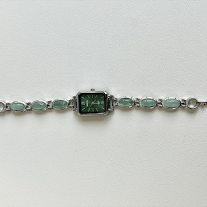 May include: Silver-toned wristwatch with a rectangular green face and matching green oval-shaped stones along the band. The watch face has silver hands and markers. The band is approximately 7 inches long.