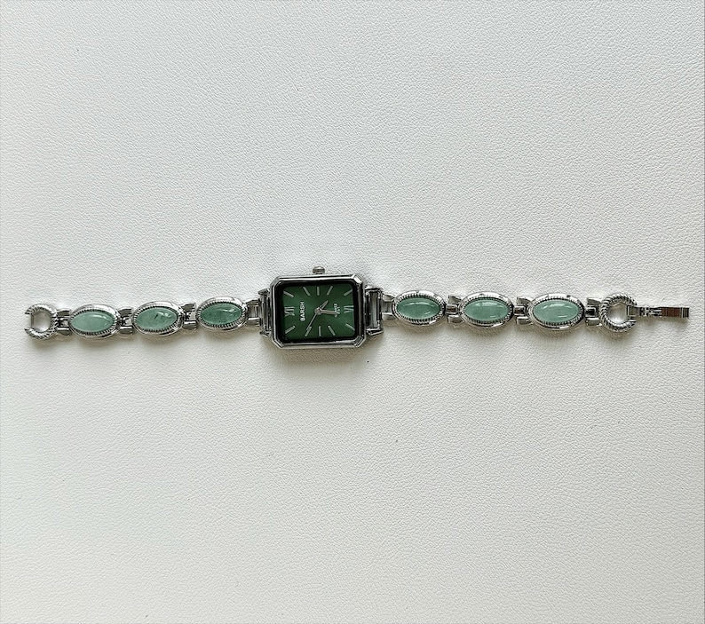 May include: Silver-toned wristwatch with a rectangular green face and matching green oval-shaped stones along the band. The watch face has silver hands and markers. The band is approximately 7 inches long.