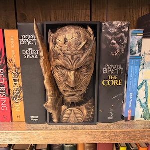 May include: A bronze-toned sculpture of a fantasy creature's face and a blade, positioned between books on a wooden shelf. The books include titles such as "Morning Star," "Red Rising," and "The Core."