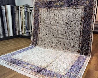 May include: Large, rectangular Persian rug with a cream-colored field and intricate floral patterns. The border is a deep navy blue with ornate designs and a cream fringe. The rug is displayed on a wooden surface.