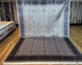 May include: A large, rectangular rug with an intricate floral pattern. The rug features a cream-colored central field with a repeating design, framed by a dark border with blue and cream accents. The rug has a fringe.
