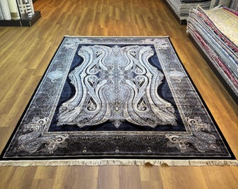 May include: A rectangular, dark blue rug with an ornate, symmetrical design in shades of white and gray. The rug features a central paisley-like pattern and a detailed border. The rug is displayed on a wooden floor in a carpet store.