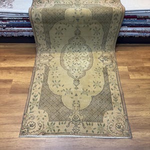 Puede incluir: Una alfombra de pasillo beige con un patrón floral verde y marrón. La alfombra tiene un diseño tradicional con un medallón central y un borde.